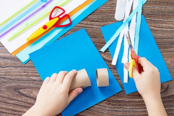 The child cuts out the details of a paper boat, congratulations concept of Father’s Day. Glue, scissors and paper on a wooden table. Children’s art project craft for kids. Craft for children.